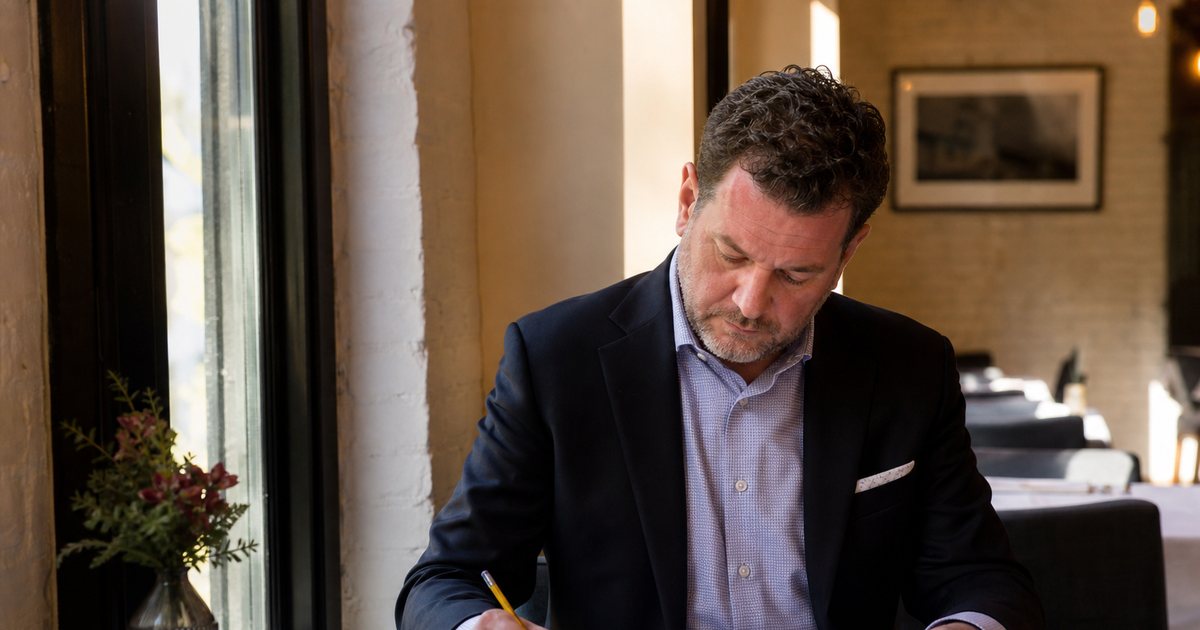 A restaurant general manager at a corner table in a quiet dining room mid-afternoon, a paper reservation ledger open beside a coffee cup, making tally marks with a pencil, sunlight through a tall window