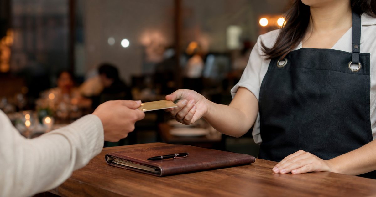 Close-up of a guest handing a credit card across a wooden restaurant host stand to a host wearing a dark apron, a leather reservation book and a pen on the counter, softly lit dining room out of focus behind