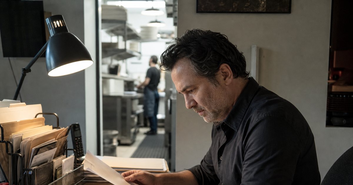 A restaurant owner at a small back-office desk stacked with paper folders and a printed reservation log, reading a letter under a desk lamp, a kitchen pass visible through the doorway, midday