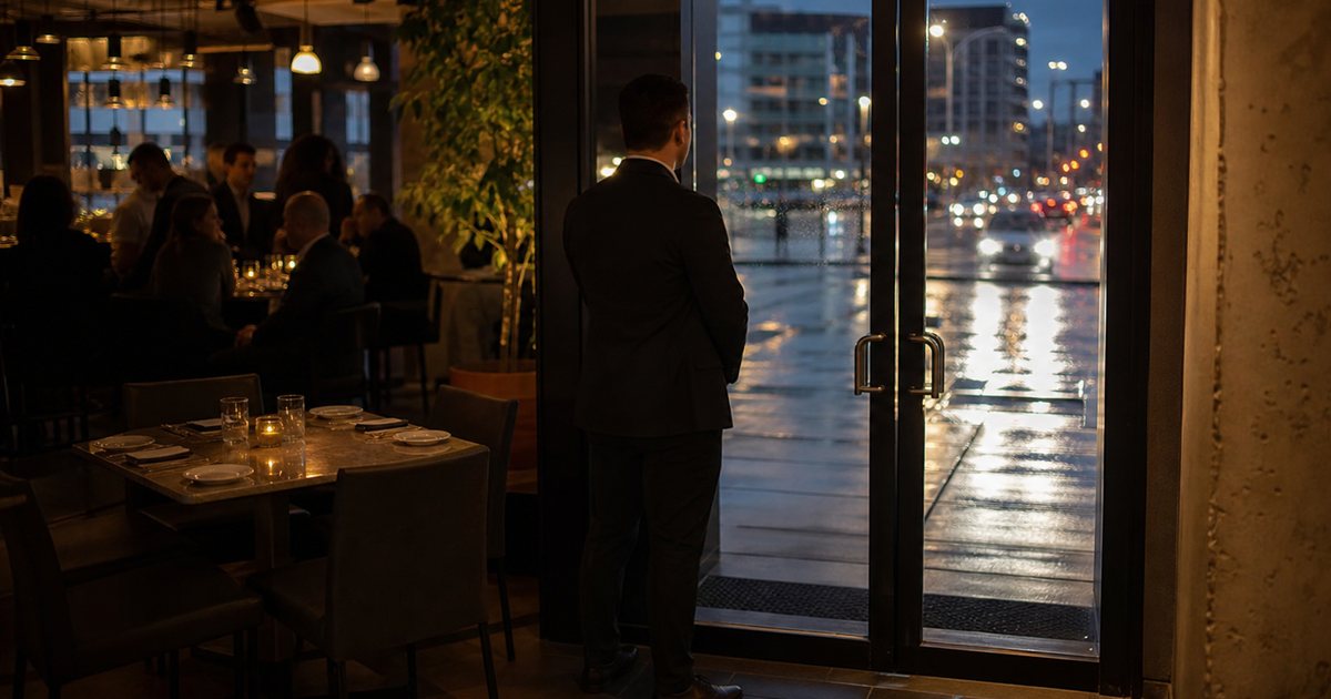 A host at a restaurant entrance looking out through the glass front door onto a rainy city street at dusk, an empty set four-top with a small candle visible in the dining room behind, guests dining at other tables