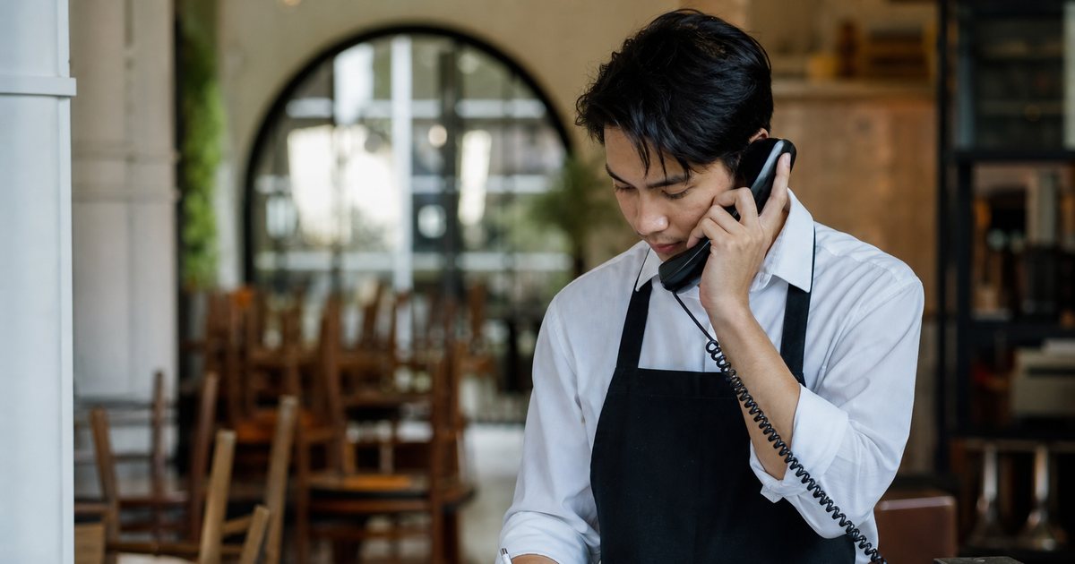 A young restaurant host in a white shirt and black apron standing at a wooden host stand with a phone handset to their ear, writing in a paper reservation book, afternoon light, empty dining room with stacked chairs behind